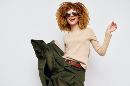 Smiling young woman in stylish outfit dancing with joy, wearing sunglasses, vibrant curly hair, standing against a light gray background, showcasing fashion and happinessの写真素材