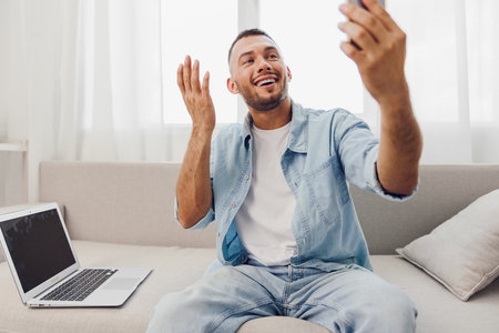 Smiling man engaged in a video call, expressing joy. Bright and airy living room setting, showcasing...