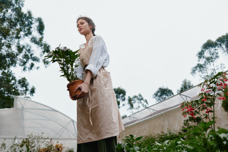 Young woman holding a potted plant in a greenhouse, wearing a casual outfit and an apron, with a serene expression reflecting her passion for gardeningの写真素材