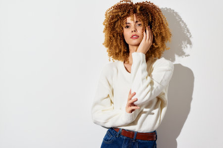 Portrait of a stylish young woman with curly hair in a cozy white sweater against a bright white background, showcasing a modern and minimalistic fashion conceptの写真素材