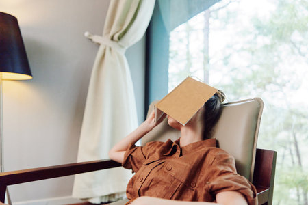 A young woman relaxing in a modern living room with a book covering her face, conveying a sense of tranquility and escape in a stylish environmentの写真素材