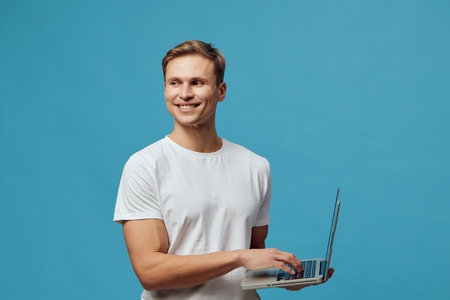 Smiling young caucasian man holding laptop wearing white t-shirt looking aside on blue background...