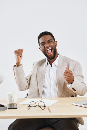 Joyful young African-American man wearing a beige blazer and white shirt sitting at office desk with...