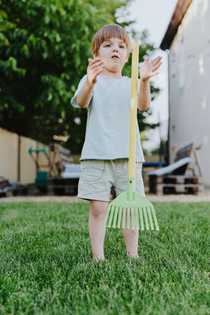 Charming boy playing outdoors with a colorful rake, enjoying a sunny day, while expressing curiosity and joy in a vibrant green garden.の写真素材