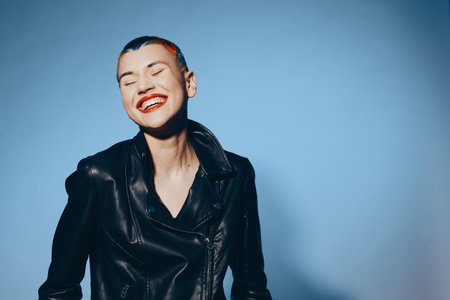 Smiling woman with short hair wearing a black leather jacket standing against a blue background,...