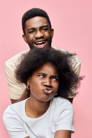 Joyful couple laughing together with playful expressions against a pastel pink background,...