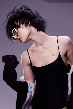 Slim woman in black dress posing with artistic hairstyle and earring against soft purple background
