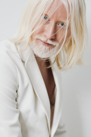 Portrait of a stylish individual with long, light colored hair and a beard, dressed in a white...
