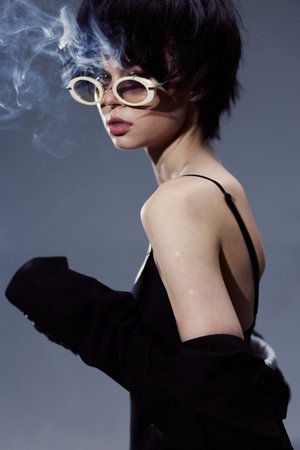 Stylish woman wearing sunglasses with smoke background, dark outfit, and short hair in moody...