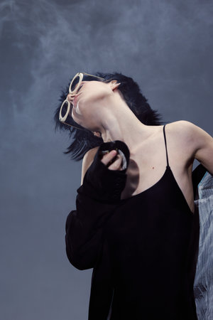 Stylish woman with short hair wearing sunglasses and black outfit posing against smoky background.の写真素材