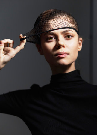 Confident woman in a stylish black outfit adjusting a netted headpiece, showcasing modern fashion and elegance against a softly lit background.の写真素材