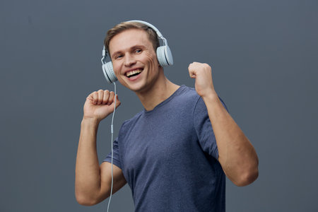 Smiling man enjoying music with headphones, showing enthusiasm and happiness, casual blue T-shirt, studio shot on solid gray background, lifestyle and entertainment concept, youthful energy, relaxation.の写真素材