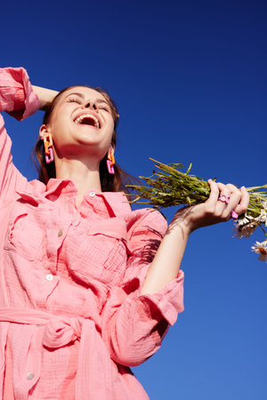 Joyful young woman in pink dress laughing while holding wildflowers against a bright blue sky, embodying happiness and nature in springの写真素材