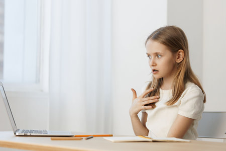 A young girl showing surprise and curiosity while studying at a desk, wearing a white shirt, in a bright and airy room with soft natural lightの写真素材