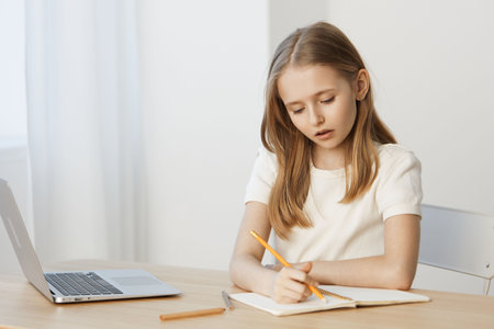 Thoughtful young girl writing in a notebook at a desk, captured in a bright and minimalistic workspace with a laptop, reflecting focus and concentration on her schoolworkの写真素材