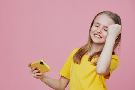 Happy girl celebrating success with a smartphone, wearing a yellow t shirt against a pink background, showcasing joy and excitement in a vibrant and playful atmosphereの写真素材
