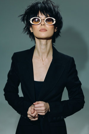 Stylish woman with short black hair wearing white round sunglasses and a black blazer posing confidently against a muted background.の写真素材
