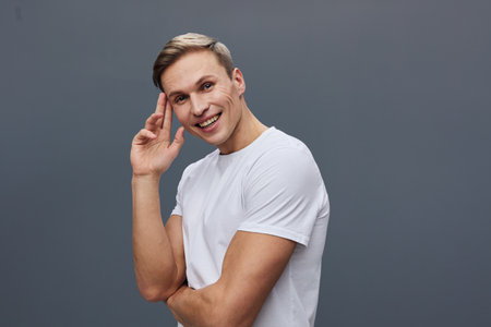Smiling young man with short blond hair wearing casual white t-shirt, looking confidently at camera, hand near head, cheerful expression isolated on plain gray background, studio portrait, people lifestyle conceptの写真素材