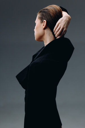 Woman in black outfit posing thoughtfully with hand behind head against gray background, elegant and minimalistic styleの写真素材