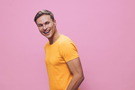 Bright cheerful young man with fair skin, short blond hair wearing casual yellow t-shirt smiles happily, standing against a pink background. Studio portrait, positive mood, lifestyle concept, vibrant colors, summer attire.の写真素材