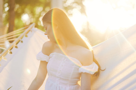 Young woman in a white dress enjoying sunlight, with a dreamy expression, surrounded by greenery and soft light reflections in spring seasonの写真素材