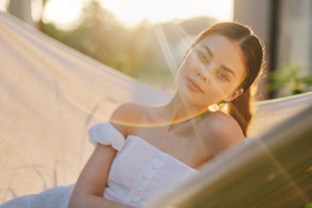 A serene young woman enjoying the golden hour in a hammock, dressed in a white off shoulder dress, radiating calmness and happiness in a lush outdoor settingの写真素材