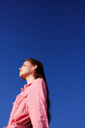 A young woman of European descent wearing a pink shirt against a clear blue sky, showcasing confidence and tranquility in a summer settingの写真素材