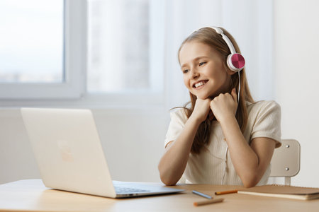 Young girl with headphones smiling at a laptop in a bright room, expressing joy and excitement while...