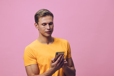 Young man in yellow t-shirt using smartphone against pink background, casual clothing, focused expression, modern technology, lifestyle concept, isolated studio shot.の写真素材