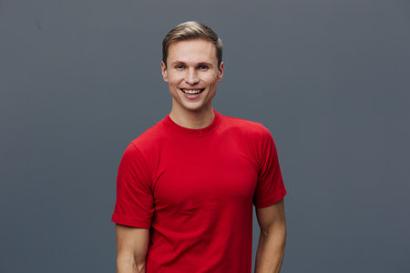 Smiling young man with short brown hair wearing a bright red t-shirt looking at camera with a cheerful expression against plain gray background. People lifestyle conceptの写真素材