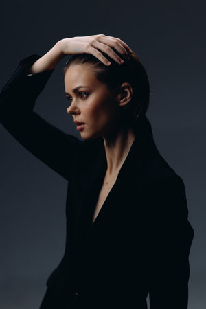 Thoughtful woman in black outfit posing with hand on head against dark background with moody lighting.の写真素材