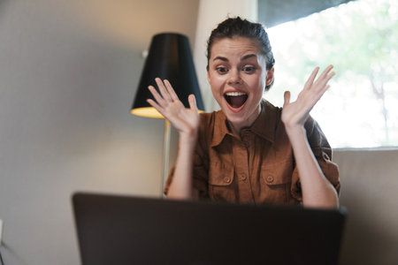 Excited young woman celebrating success at her laptop, dressed in casual brown outfit, expressing joy in a cozy home office setting with a warm color paletteの写真素材