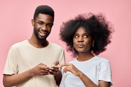 Young Black couple with stylish hairstyles, smiling and using smartphone against a pink background, representing modern relationships and technologyの写真素材