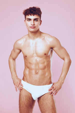 Confident young man with athletic build posing in white swimwear against a pastel pink background. Fitness and body positivity concept.の写真素材