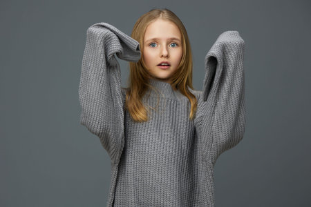 Young girl in oversized gray sweater expressing surprise with hands raised, against a neutral background, perfect for fashion and seasonal conceptsの写真素材