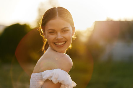 Happy young woman smiling outdoors during sunset, wearing a white dress, in a serene natural setting, exuding joy and warmthの写真素材