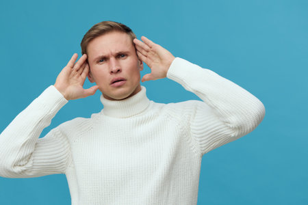 Frustrated young caucasian man wear white sweater hold hands near head face stressed emotion isolated blue background mental health conceptの写真素材