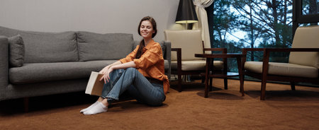 Young woman in casual attire sitting on the floor, smiling with a book in a cozy living room,...