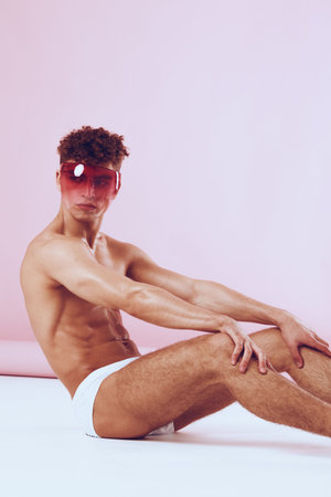 Stylish young man sitting on the floor in minimalistic pose, wearing white briefs and a colorful visor. Vibrant fashion and confidence concept.の写真素材