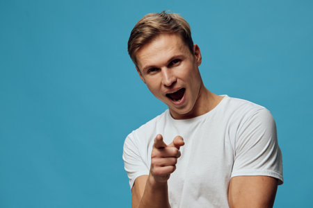 Excited young caucasian man in white t-shirt pointing finger yelling over blue background studio portrait expression conceptの写真素材