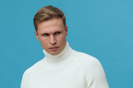Close-up of a young man with short light brown hair wearing a white knitted turtleneck sweater on...