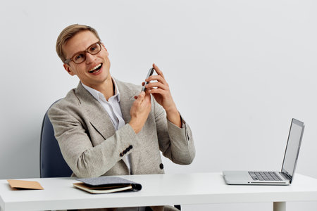 smiling man sitting at desk wearing glasses grey blazer holding smartphone looking at camera with...