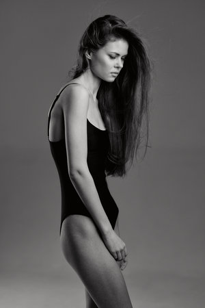 Black and white portrait of a slender woman in a leotard with long hair, expressing pensive mood in minimalistic studio setting.の写真素材