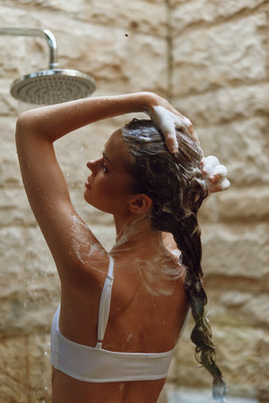 Woman washing hair in outdoor shower with natural stone background, foam lather, wet bra, and relaxed expression during warm daylight.の写真素材
