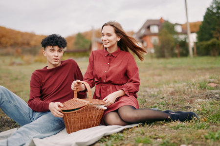 Two peers sit on autumn grass, one with a wicker basket, sharing a quiet moment in soft light. Authentic, candid interaction captures genuine warmth and calm confidence.の写真素材