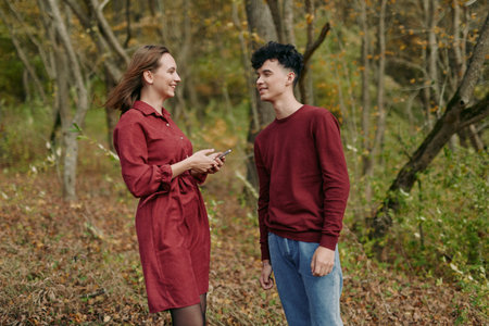 Young woman and teenage boy converse in a wooded clearing, smiling and holding a phone, natural...