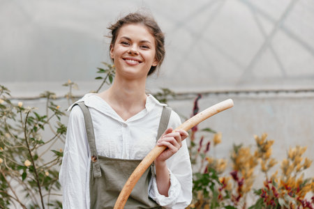 Happy young woman in a green garden holding a wooden stick, wearing casual grey overalls and a white shirt, emphasizing a cheerful, eco friendly lifestyleの写真素材