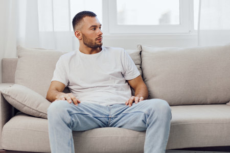 Thoughtful man sitting on a cozy sofa, reflecting in a serene indoor environment with natural light. Stylish outfit complements modern decor and calm atmosphere.の写真素材