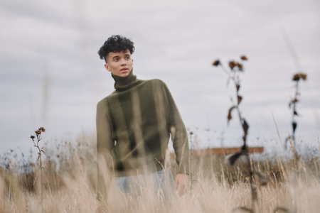 Young man in a green turtleneck stands among tall dry grasses under soft overcast light,...