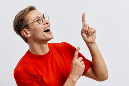 Cheerful young caucasian man wear red tshirt glasses pointing up smiling look side isolated on light...